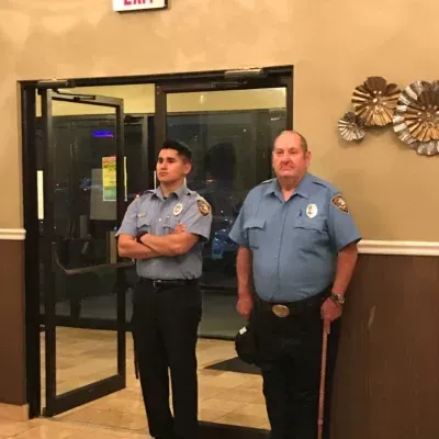 Two security guards stand at an entrance: young man with arms crossed, older man with cane.