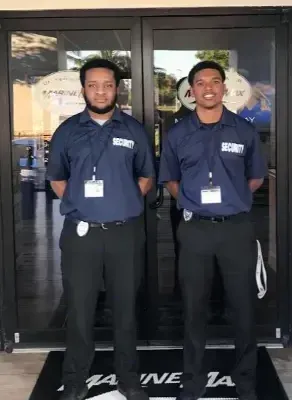 Two men in blue shirts and black pants stand in front of a store entrance.