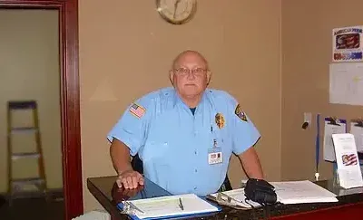 Security guard at a desk in a building, wearing a light blue uniform with patches and a name tag.