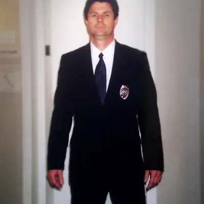 Man in a black suit with a badge, standing inside a building.