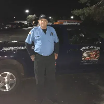 Man in uniform stands by dark blue truck, night setting. American Pride logo on truck.