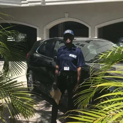 Woman in security uniform stands by a black car in front of a house, palm fronds in foreground.