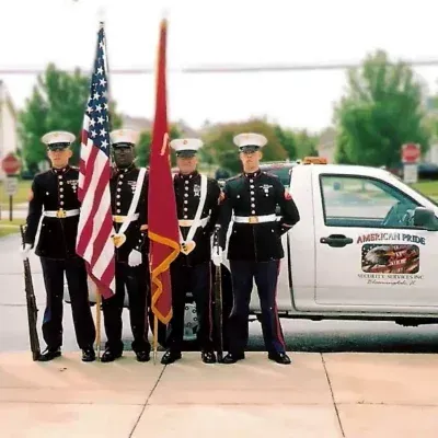 Four Marines in uniform with flags, standing in front of a white truck with 