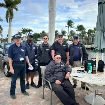 Group of men outdoors by water; some wear navy caps, jackets. One man sits at table.