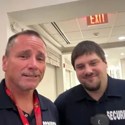 Two security guards smiling near an exit sign. One has a lanyard, the other has a radio.