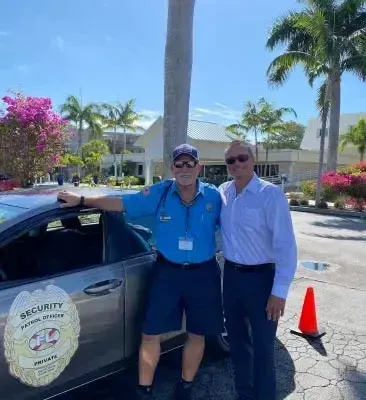 Two men pose by a security car; one in uniform, one in business attire. Outdoors, sunny day.