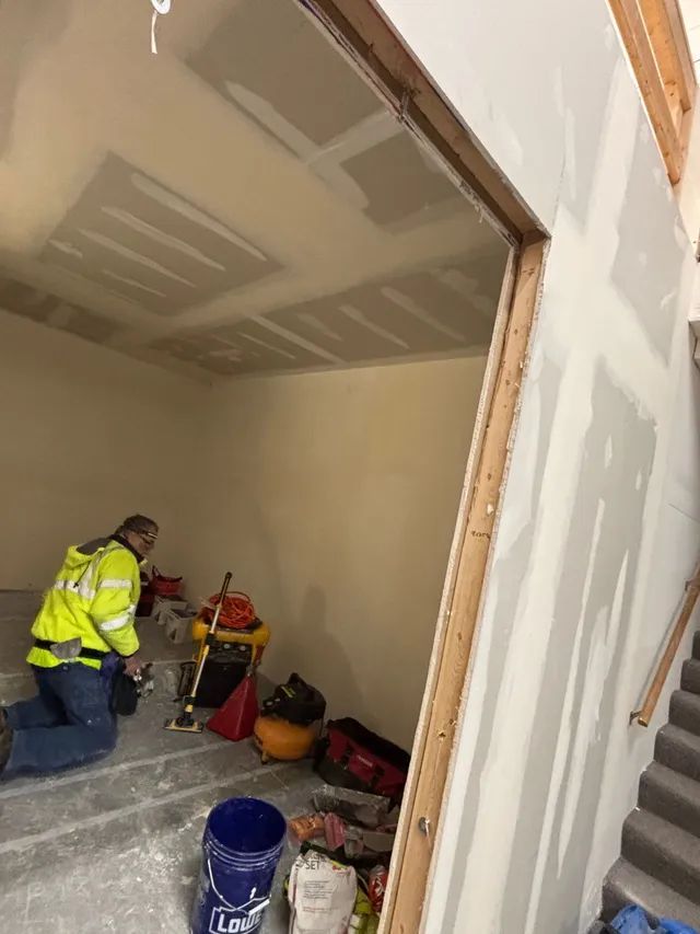 A man in a yellow jacket is kneeling in a room next to stairs.