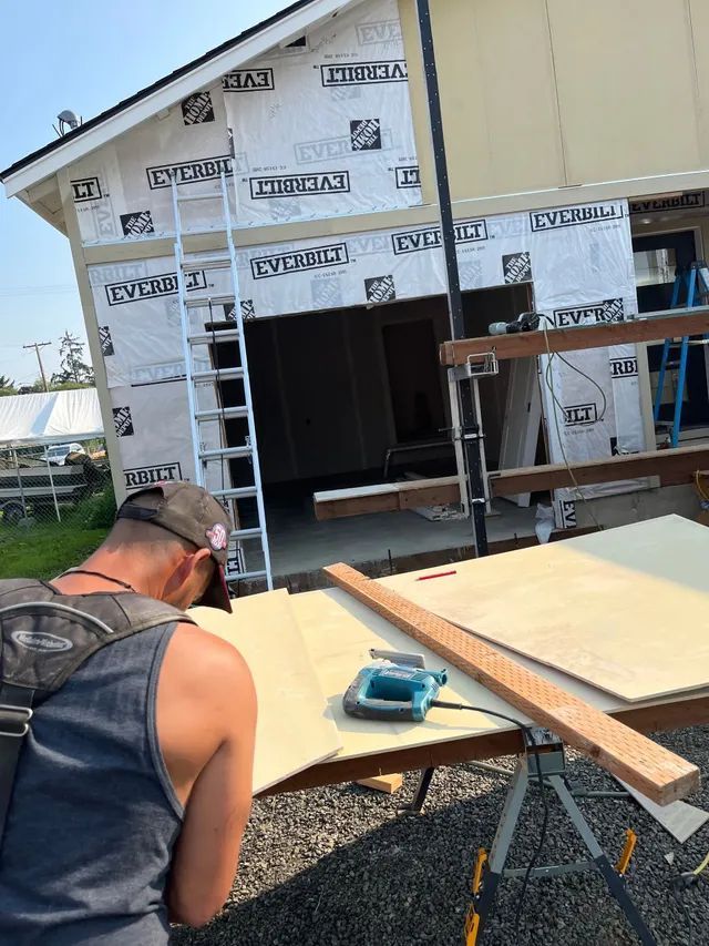 A man is working on a piece of wood in front of a house under construction.