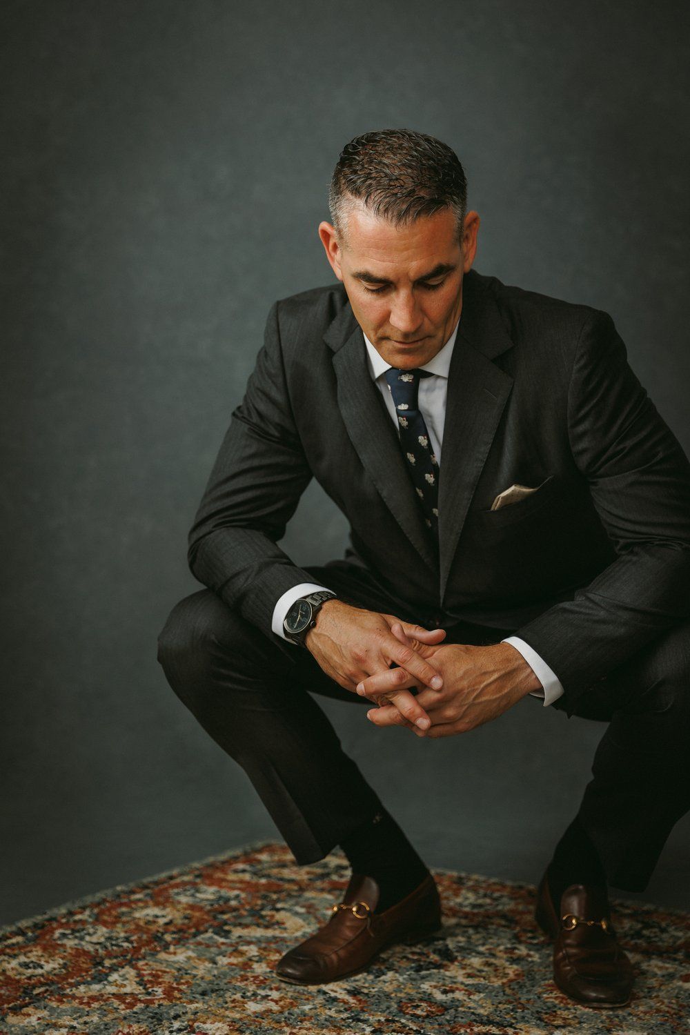 A man in a suit and tie is squatting down on a rug.