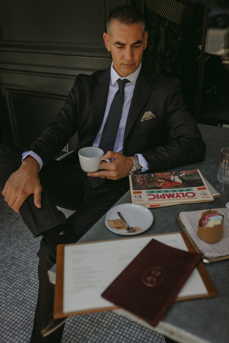 A man in a suit and tie is sitting at a table holding a cup of coffee.