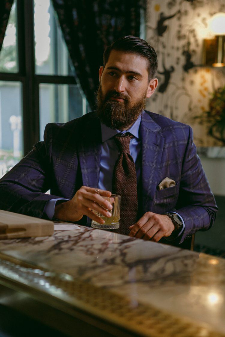 A man in a suit and tie is sitting at a bar holding a glass of whiskey.