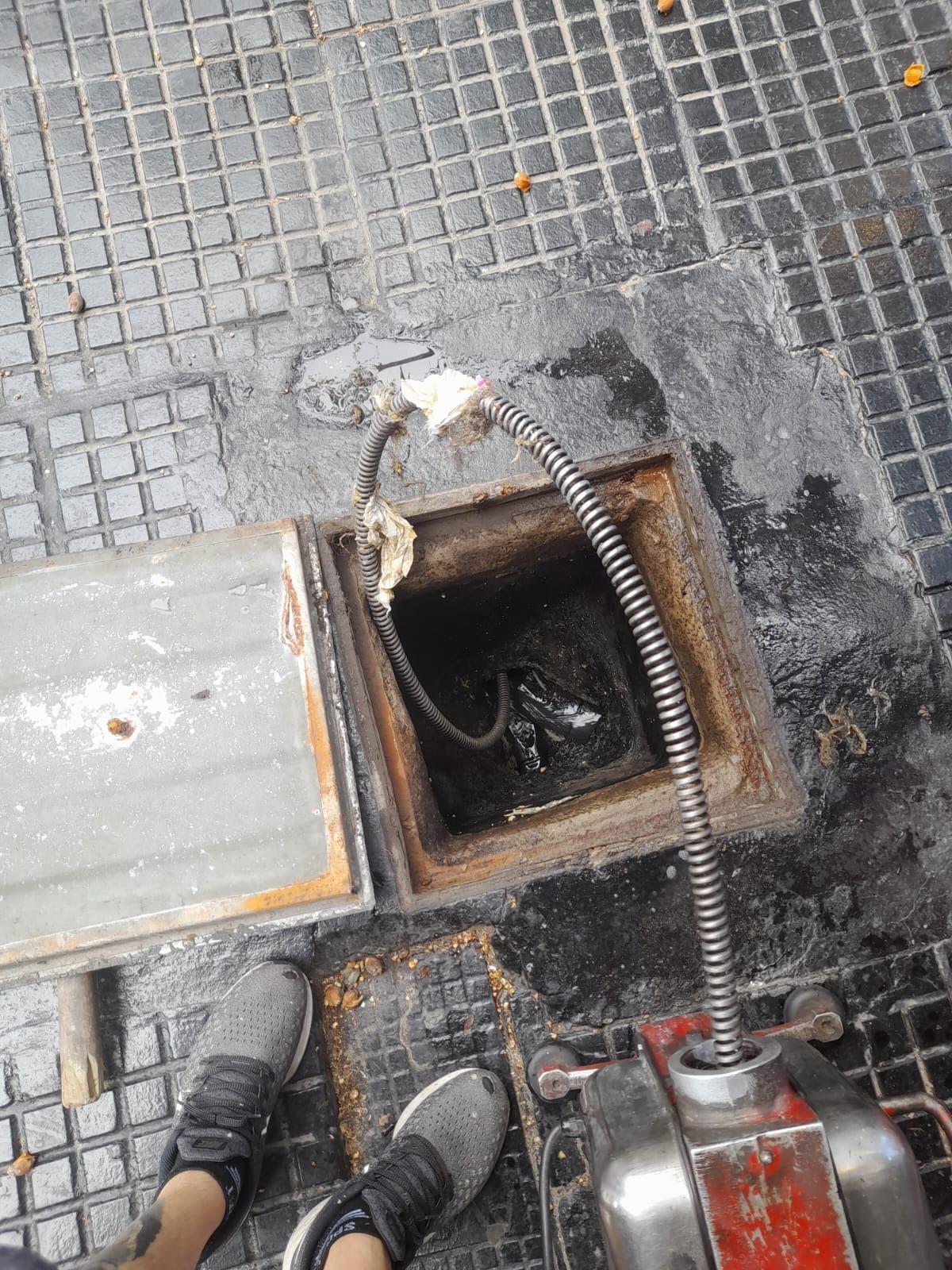 Desagüe público abierto con un cable o manguera que lo alimenta, junto a un bordillo gris y adoquines.