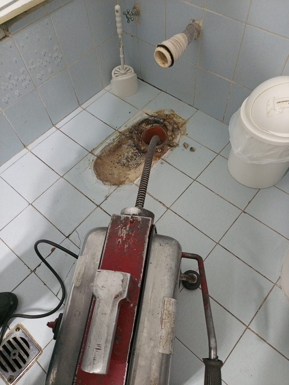 Se está limpiando un desagüe atascado del baño con una sonda desatascadora roja junto a un inodoro y un cubo.