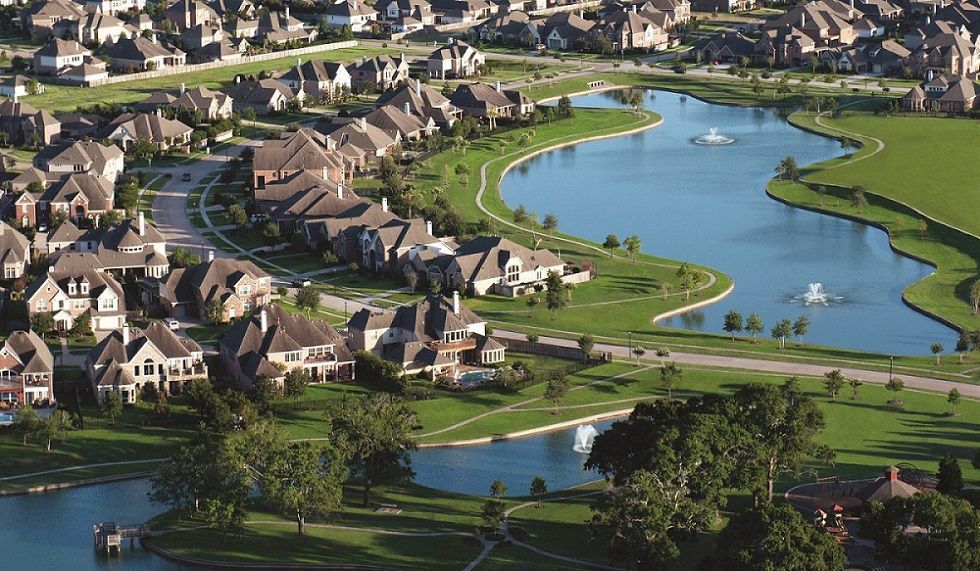 Suburban neighborhood with large houses bordering a lake with fountains and a green grassy area.