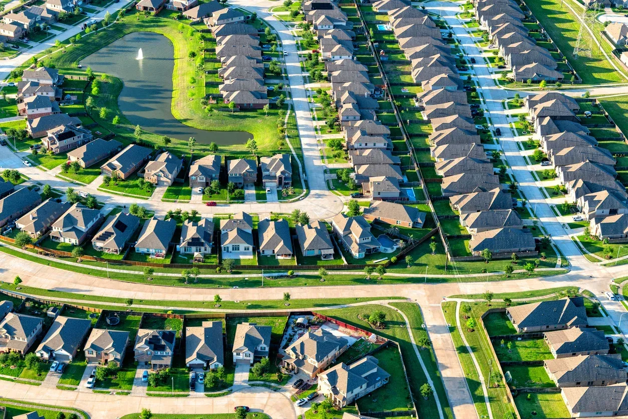 Aerial view of a suburban neighborhood with rows of houses, green lawns, a pond, and curved roads.