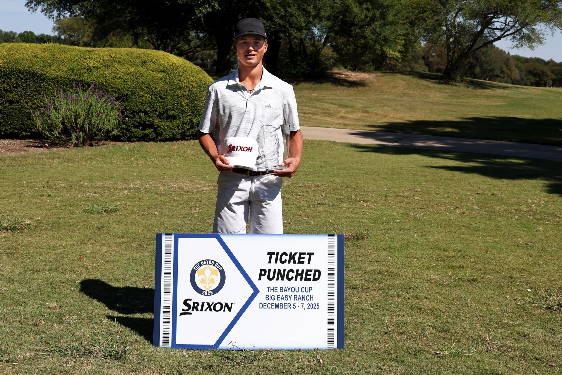 Golfer holding trophy, standing on green. He wears a pink shirt, grey shorts, and a cap. Golf bag nearby.
