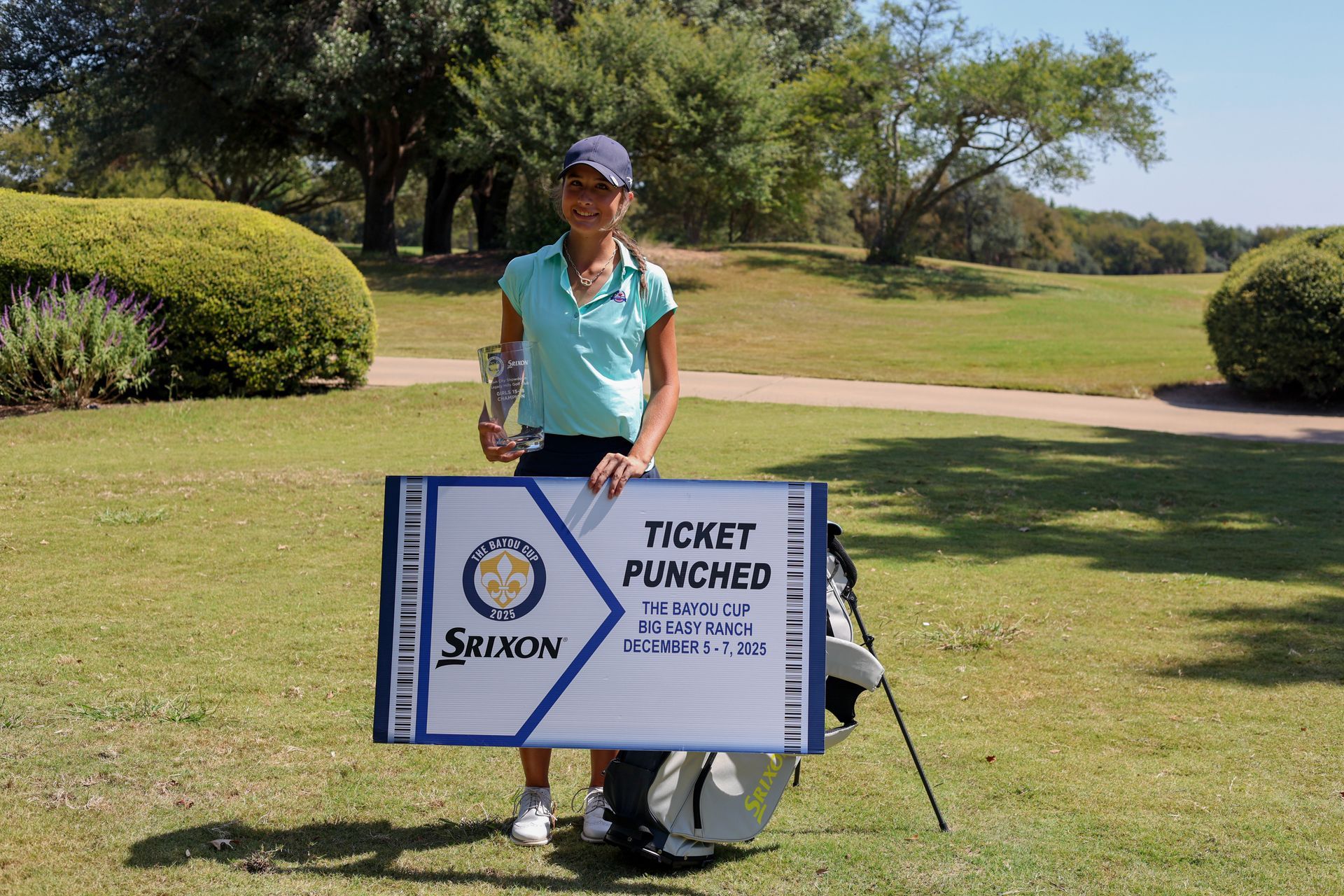 Young woman golfer in pink shirt and black shorts stands next to golf bag on a green course.