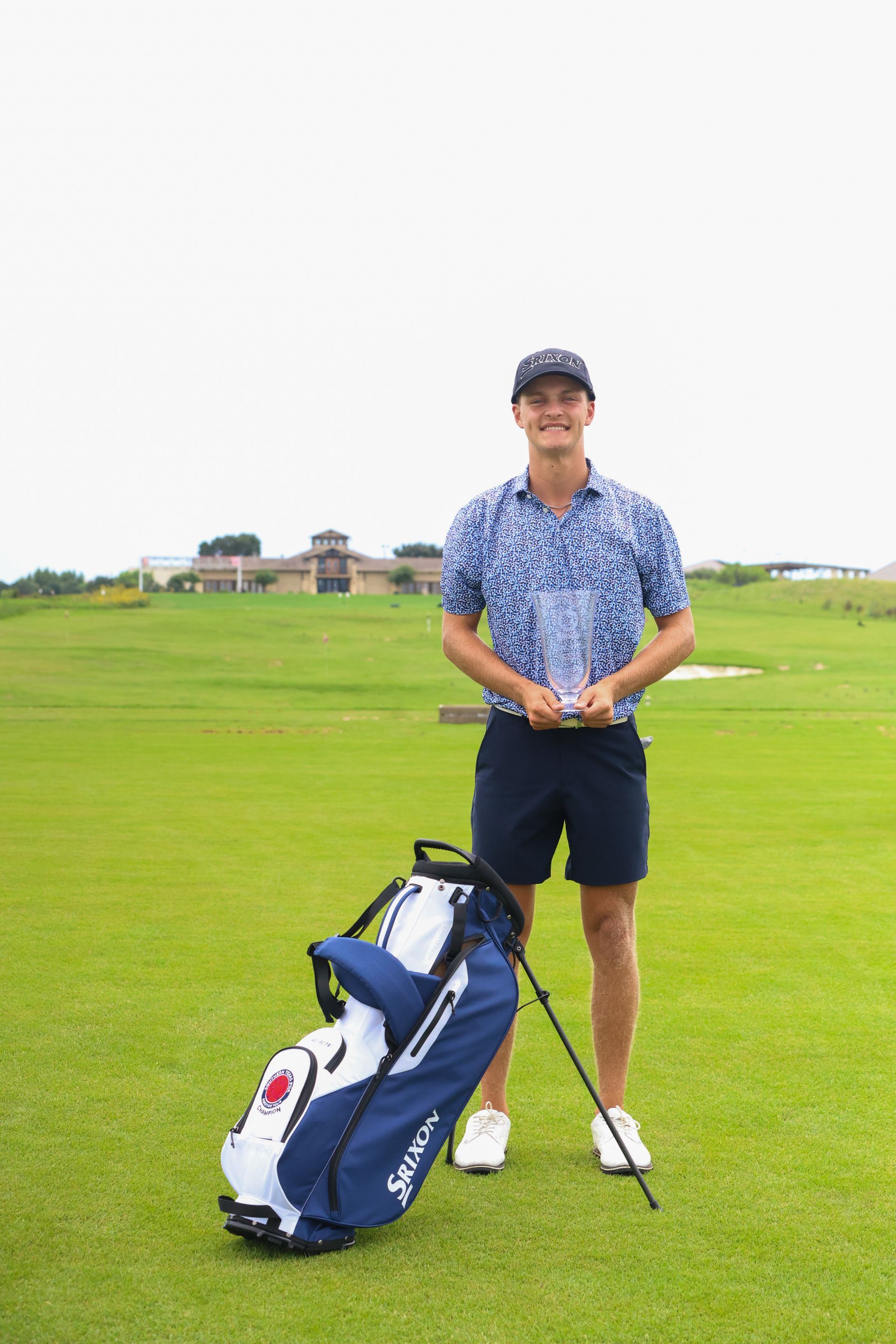 A man is standing on a golf course holding a trophy and a golf bag.