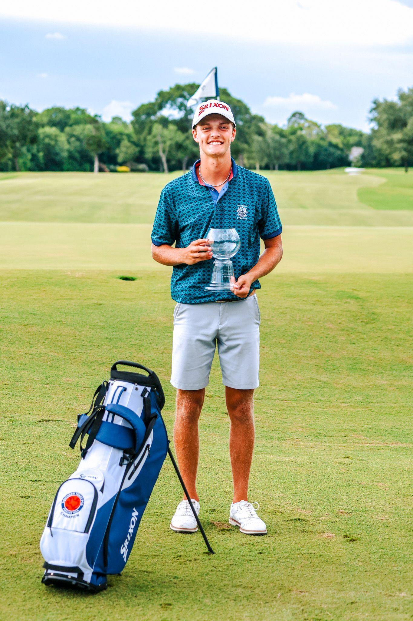 A young man is standing on a golf course holding a trophy and a golf bag.
