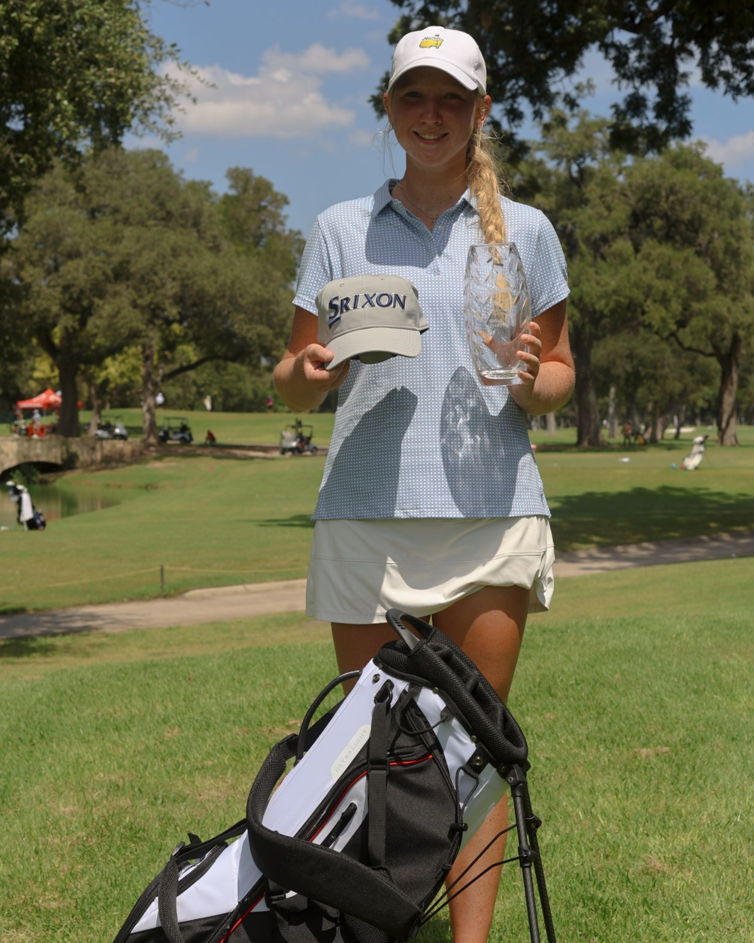 Young woman golfer in pink shirt and black shorts stands next to golf bag on a green course.