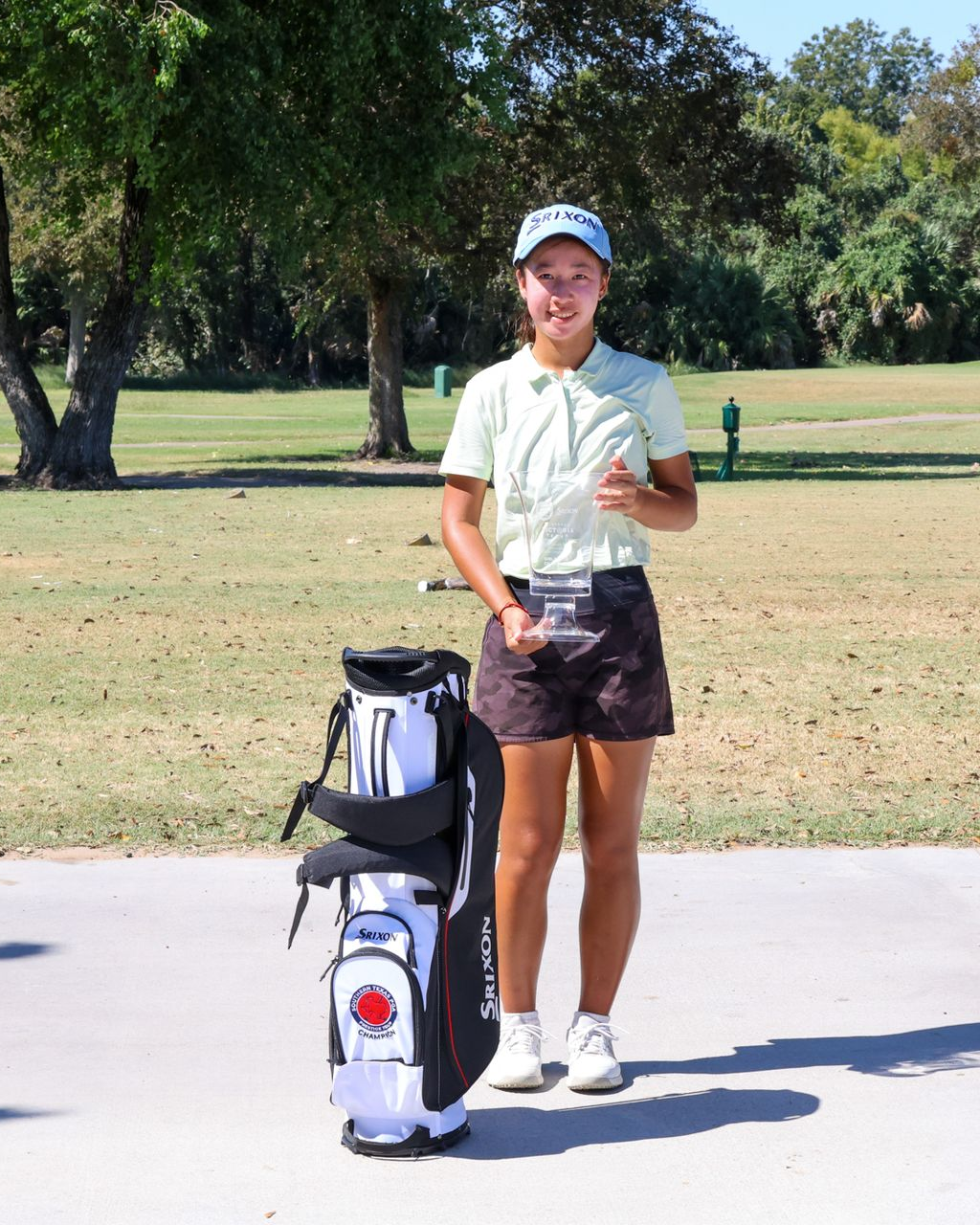 Young woman golfer in pink shirt and black shorts stands next to golf bag on a green course.