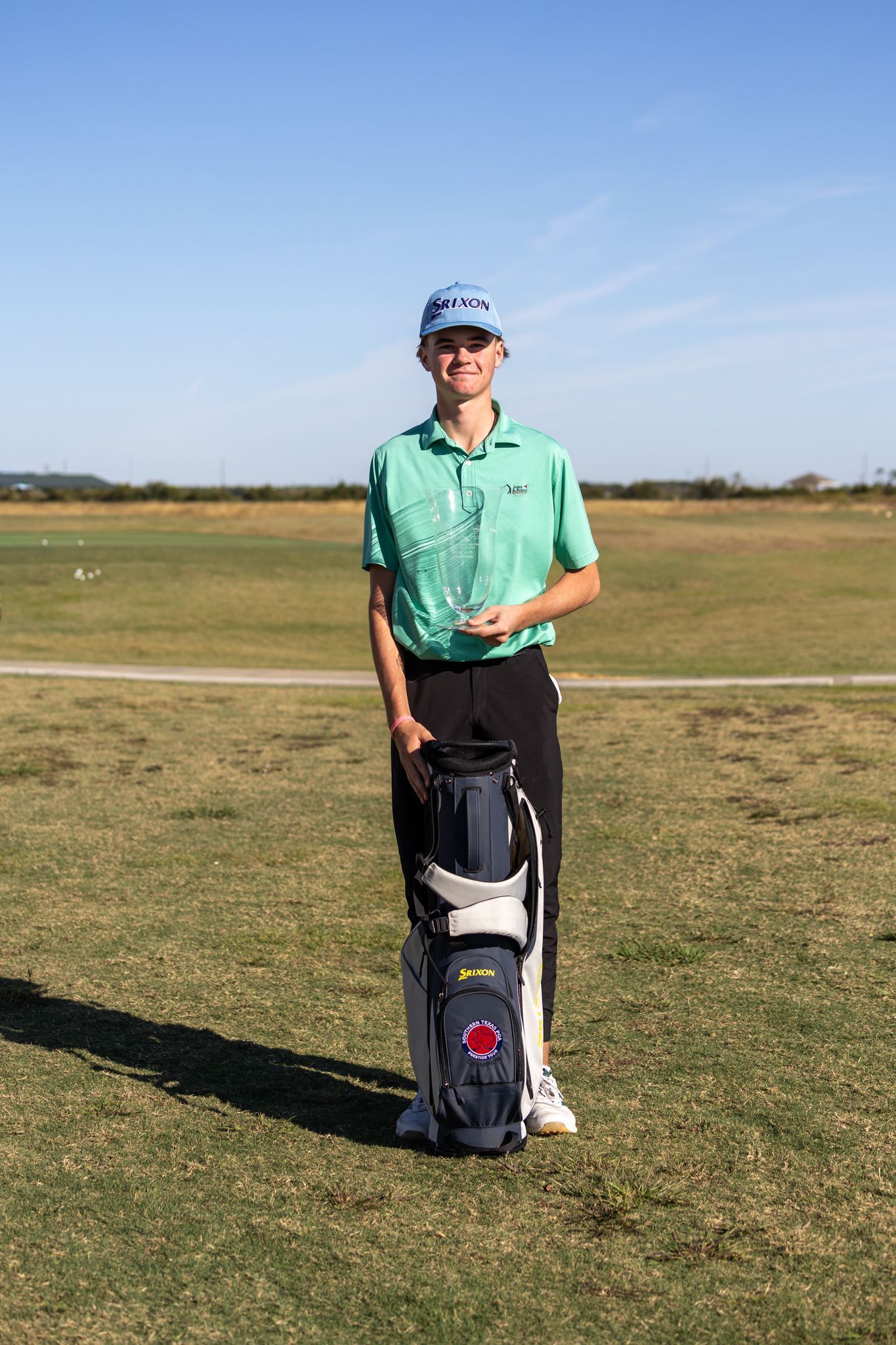 Young woman golfer in pink shirt and black shorts stands next to golf bag on a green course.
