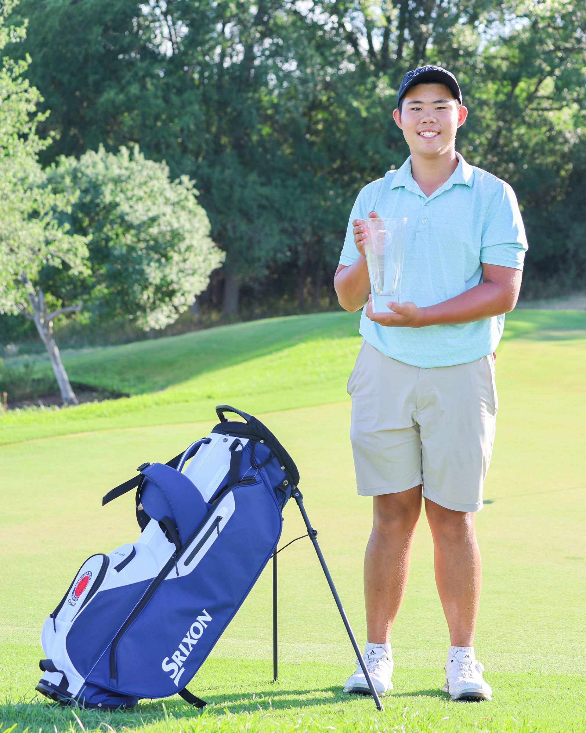 A young man is standing on a golf course holding a trophy and a golf bag.