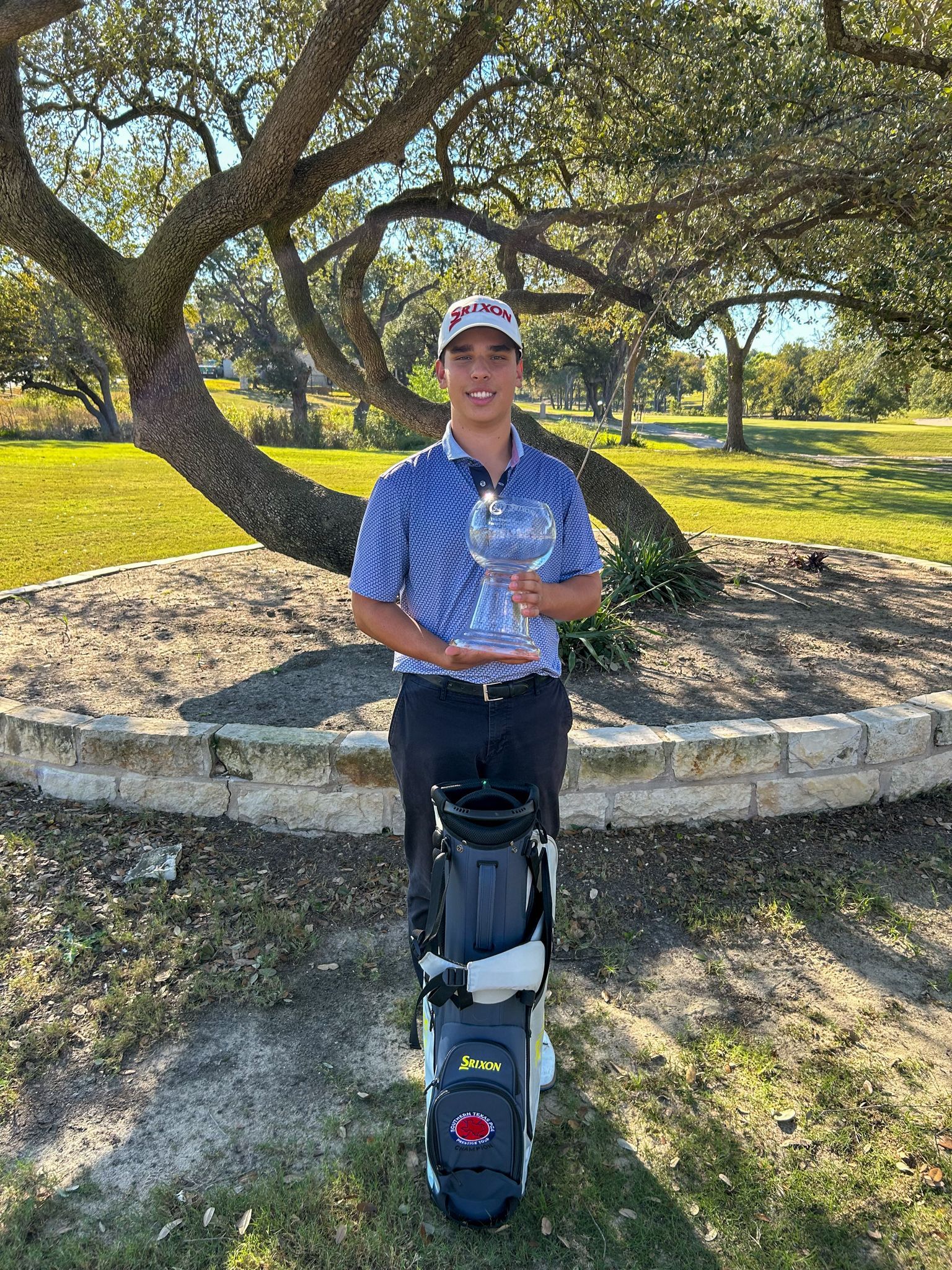 Golfer holding trophy, standing by golf bag under tree on course. Sunny day.