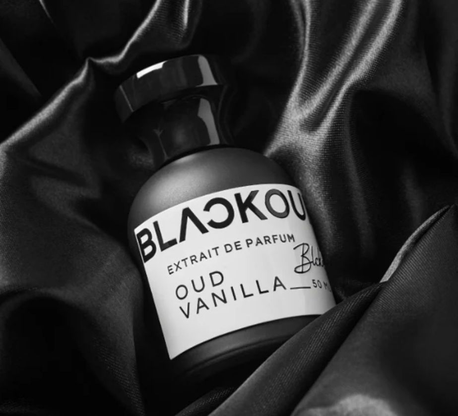 Frasco de perfume en blanco y negro, etiqueta “BLACKOU” y “OUD VANILLA”, sobre tela de satén oscuro.