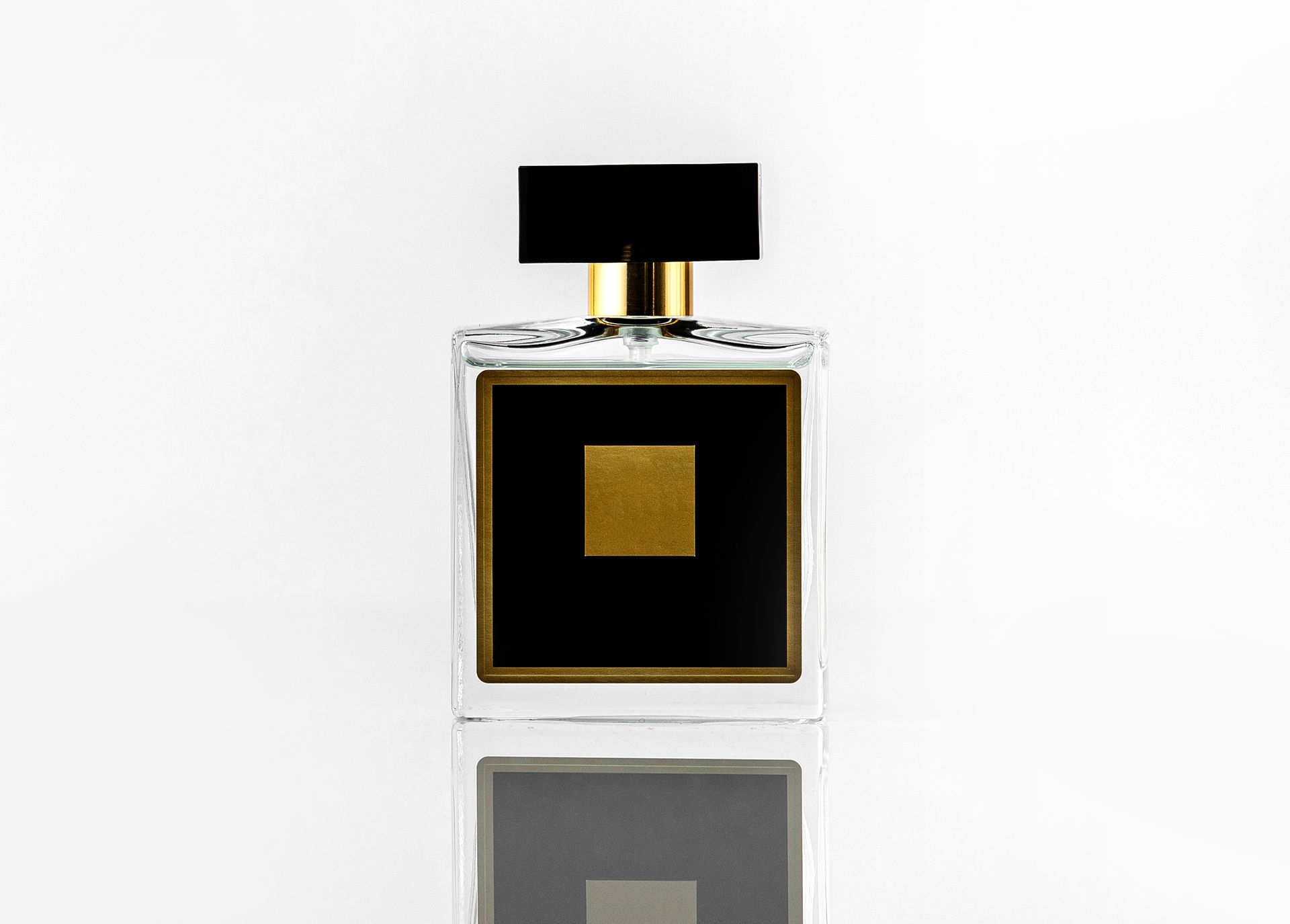 Frasco de perfume con diseño cuadrado negro y dorado sobre superficie reflectante blanca.