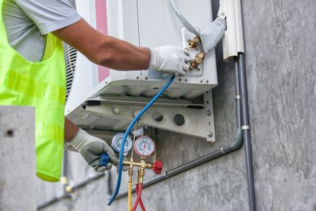 HVAC technician wearing gloves working on an AC unit, checking gauges and connections on a gray wall.