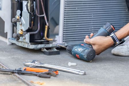 Person using a drill to repair an air conditioning unit outdoors; tools on the ground.