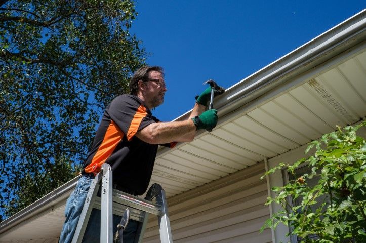 Man on a ladder cleaning gutters on a house roofline with a green tool