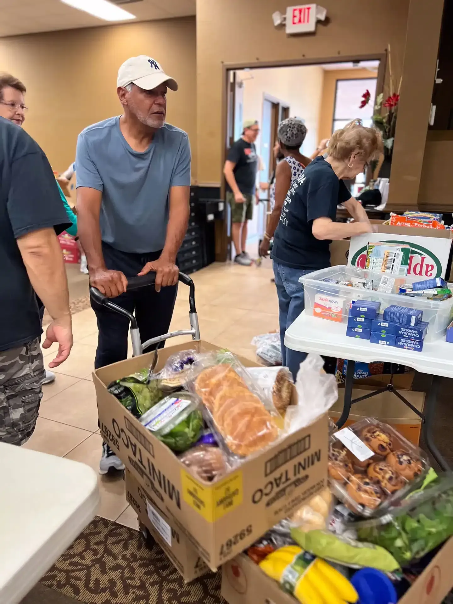 People gather at a food distribution site; a man with a walker pushes a box of groceries.