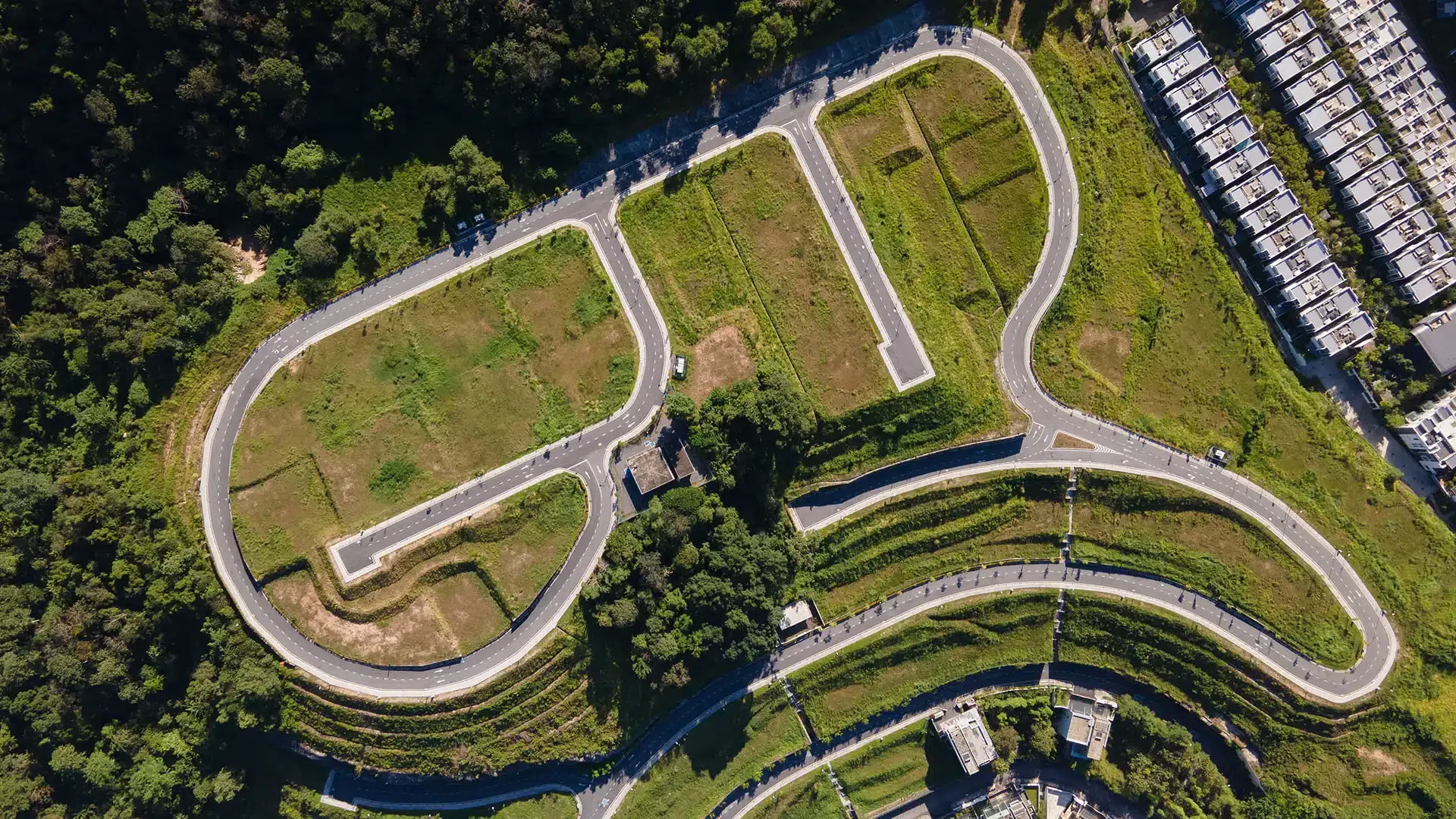 Aerial view of a road layout with partially built lots. Green grass surrounds paved roads. Residential buildings are visible on the right.