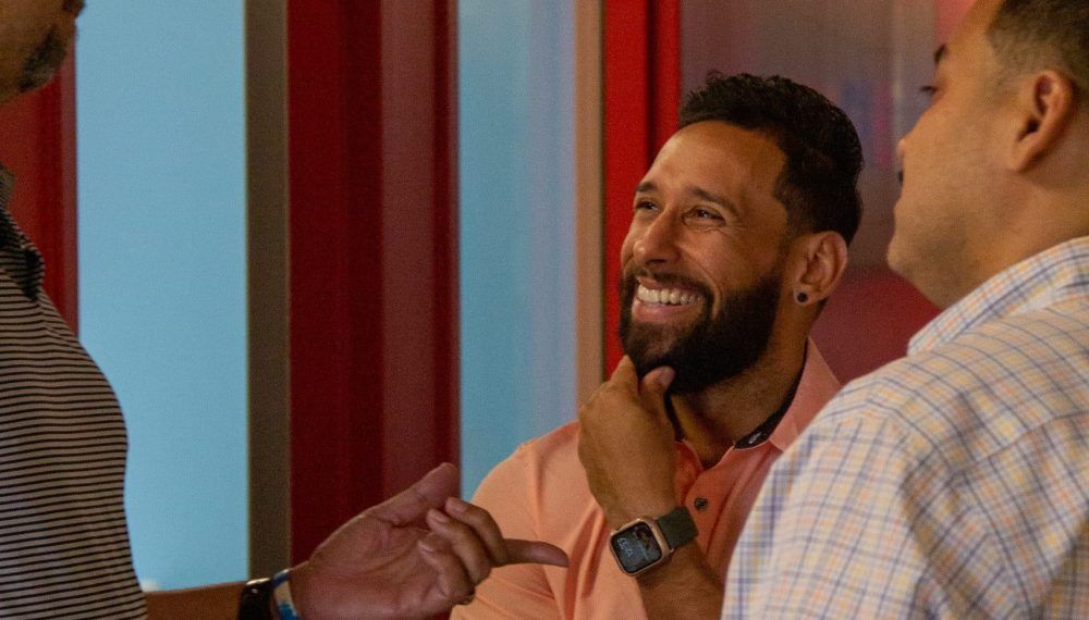 Three men in a brightly lit setting, one smiling with hand on chin, others facing him in conversation.