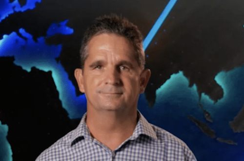Man in blue plaid shirt smiles in front of a blue world map graphic.