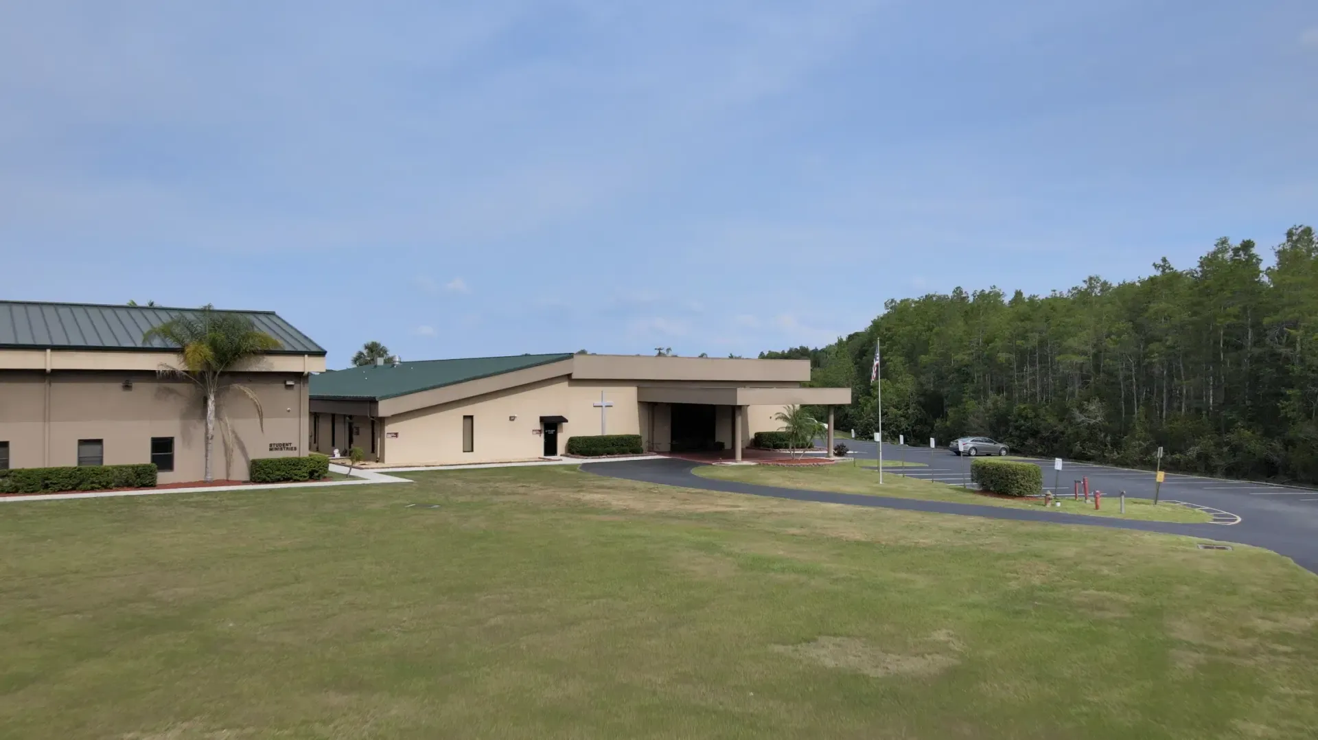 Low-angle view of a beige building with a green roof, on a grassy lawn with trees in the background under a blue sky.