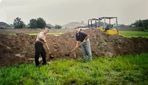 Two people raking soil in front of a dirt pile and machinery on a green grassy field.