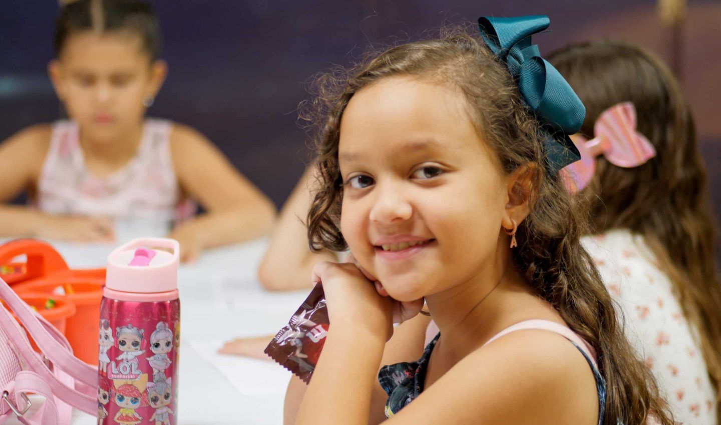 Girl with a big teal bow smiles at the camera, seated at a table with other children.