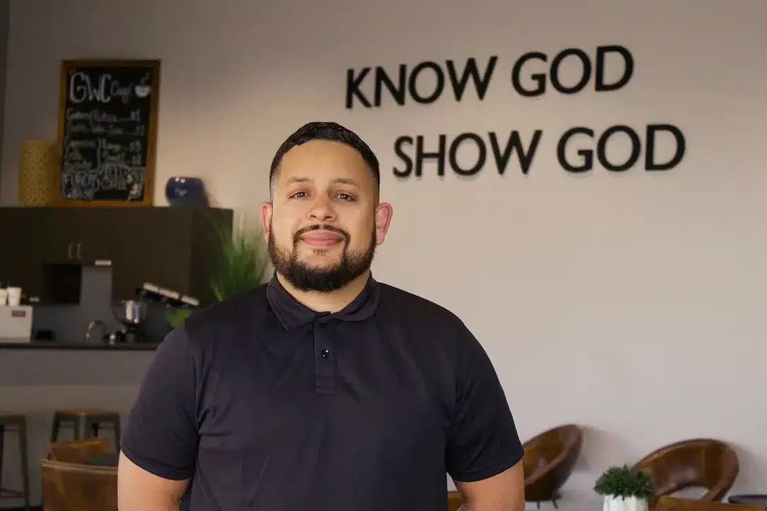 Man with a beard in a black shirt, stands in front of a wall that reads “KNOW GOD SHOW GOD.”