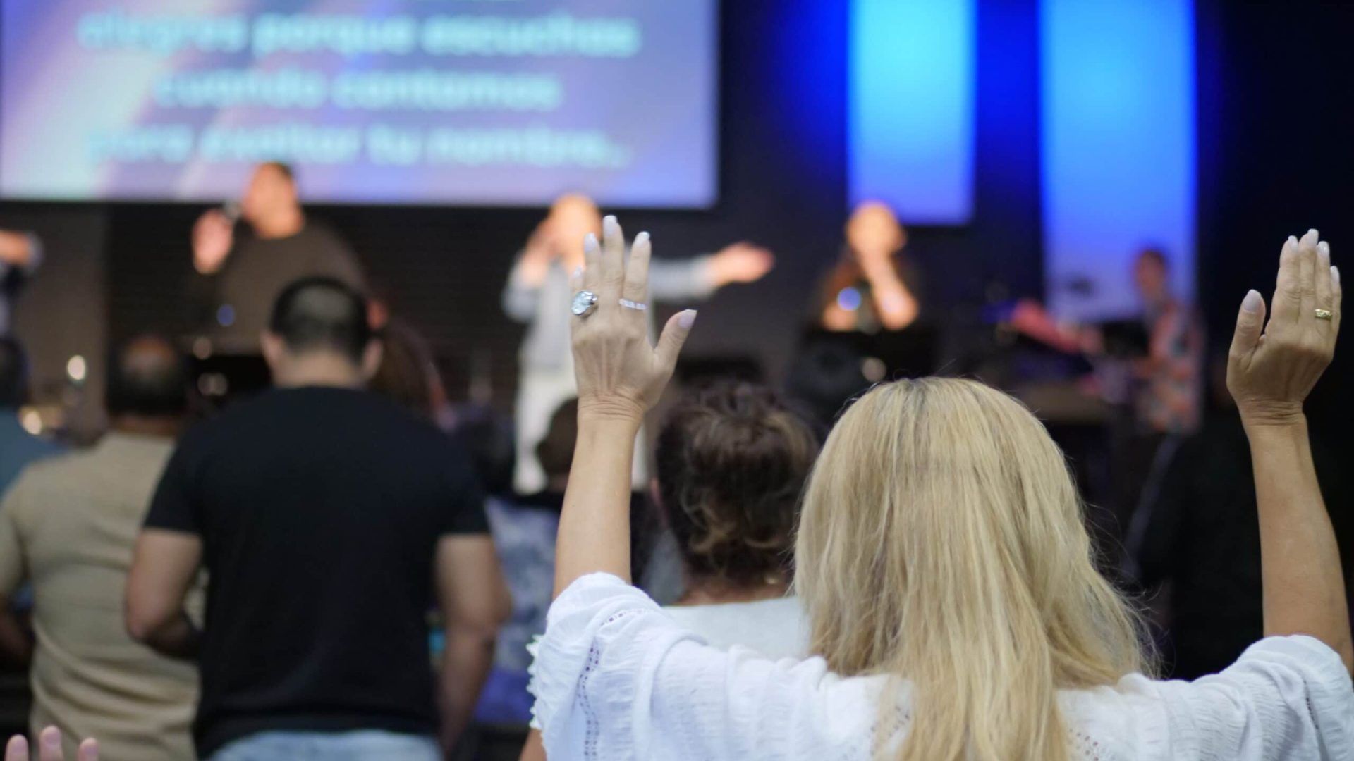 People raising hands in worship, stage with musicians and lyrics projected on screen.