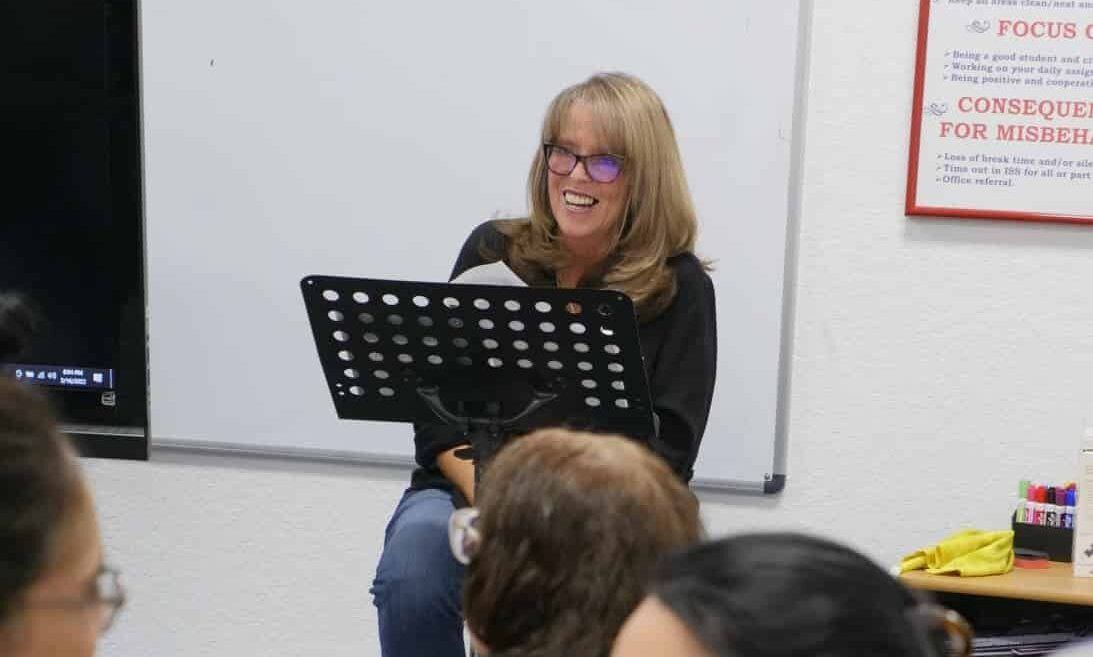 Woman with glasses smiles while reading from a music stand in a room, looking at an audience.