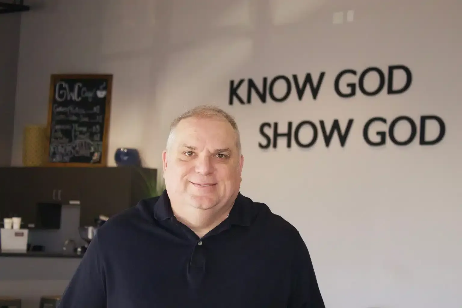 Man in a navy shirt smiles in front of a wall that says 