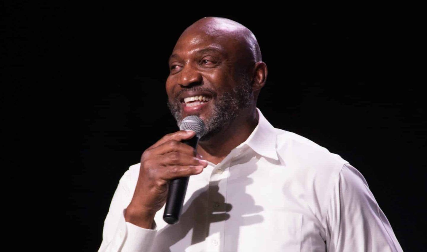 Man in white shirt holds a microphone, smiling, on a stage with a dark background.