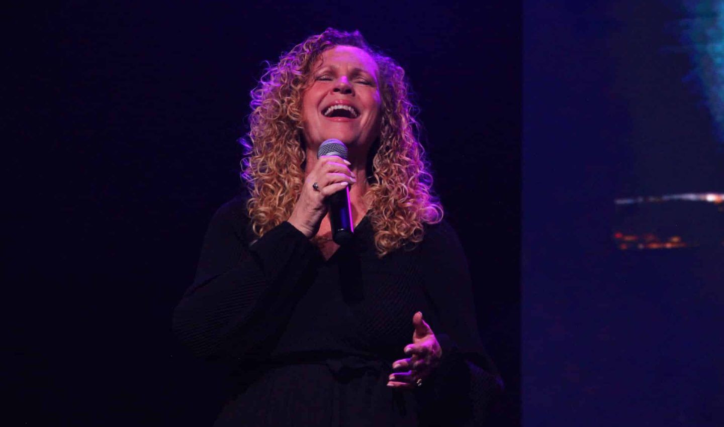 Woman with curly hair singing into a microphone on stage, smiling with arms open. Dark background.