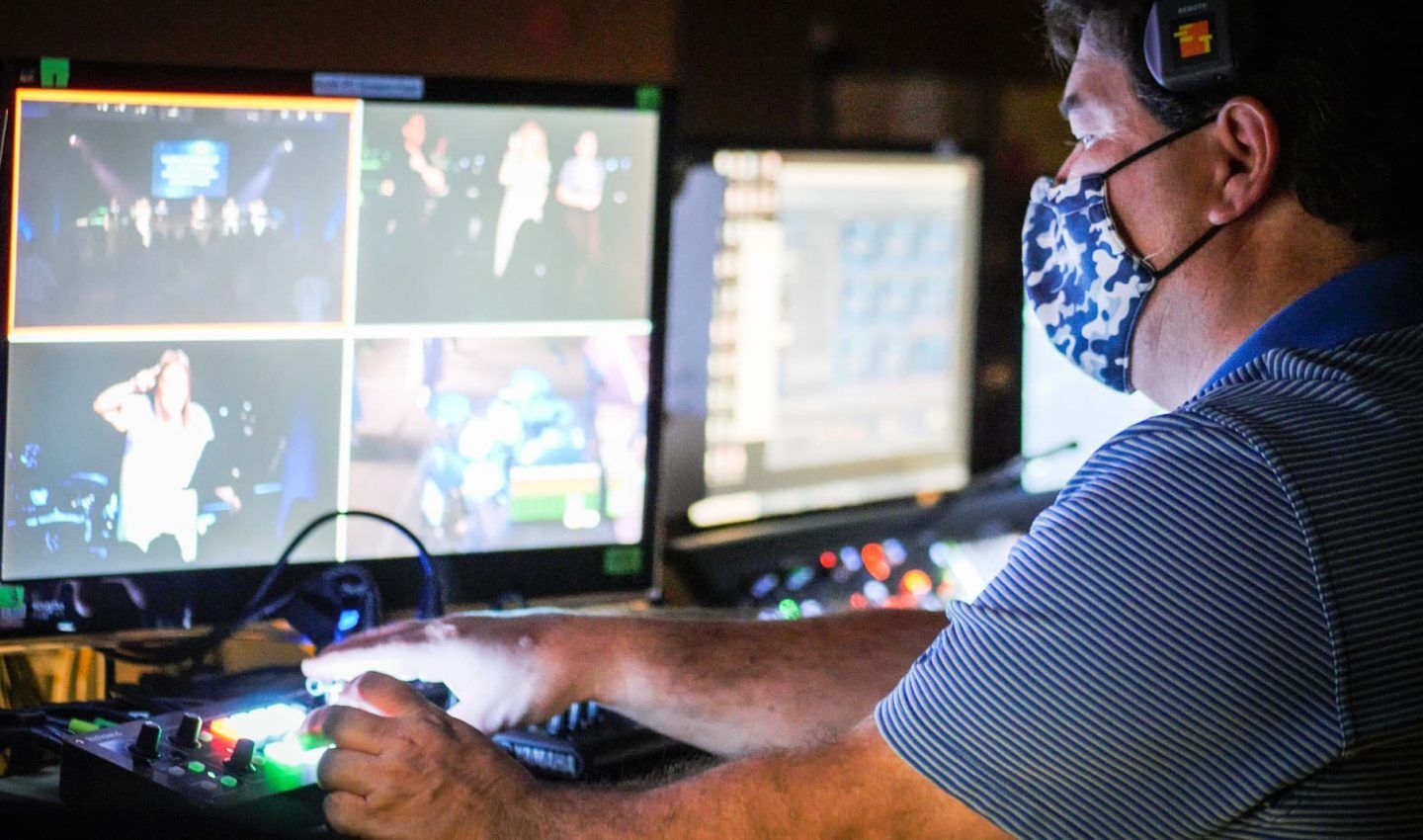 Man wearing a mask operates video equipment, watching multiple screens showing a performance.