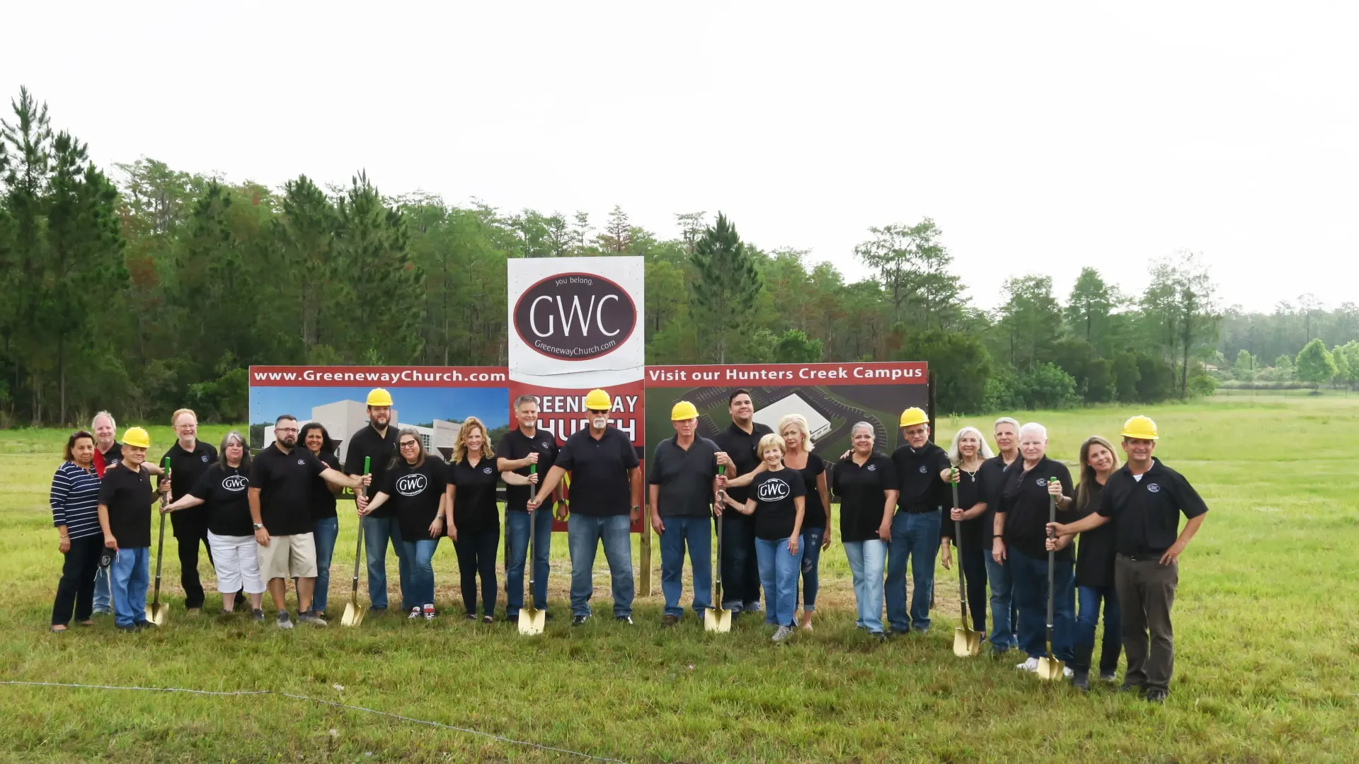 Group breaking ground with shovels, wearing hard hats, in a field with a banner that has a company logo on it.