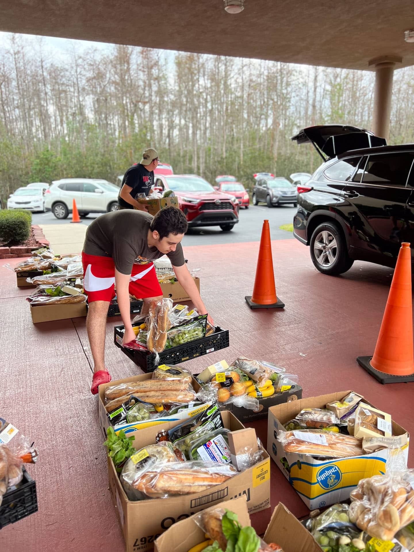 Volunteers loading groceries into cars at an outdoor distribution event. Boxes of food are on the ground.
