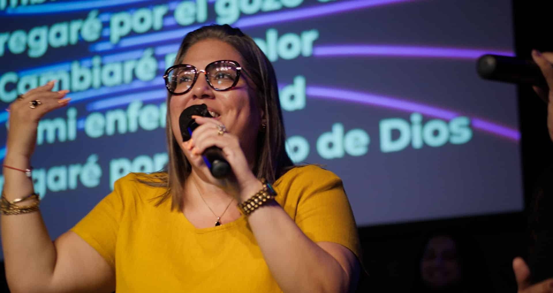 Woman singing into a microphone onstage; wearing glasses and a yellow shirt. Lyrics displayed behind her.